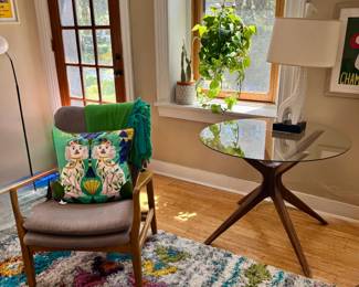 Mid-century wood-frame armchair with gray cushion, Staffordshire dog needlepoint pillow, mid-century glass-top jax/spider-leg side table, ceramic giraffe lamp base with drum shade, floor lamp, planters.