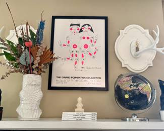 Girard Foundation Collection framed print (Museum of International Folk Art), white ceramic textured vase with dried floral arrangement, antler wall hook, vintage world globe, black cat and dog figurines, small architecture/design books (Aalto, Eames, Case Study Houses), decorative wall plaques.