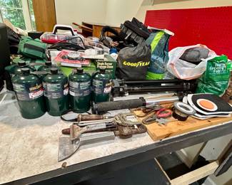 Wide assortment of camping gear, workshop tools, plumbing tools and fittings, propane stoves and fuel cylinders, Big Green Egg accessories, tripods, storage cases, organizers, hardware, utility items, and garage essentials.
