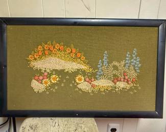 This is a vintage 1970s framed crewel embroidery wall art piece featuring a textured floral and rock garden landscape on an olive green textured background.