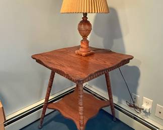 This is an antique two-tier Victorian or Edwardian parlor table, crafted from solid oak with turned spindle (or spool) legs and a lower display shelf. On top sits a vintage turned-wood table lamp with a warm, honey-toned finish and a classic pleated drum shade.