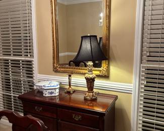 buffet features and elegant raised-panel cabinet design topped with classic black-shaded table lamp  and gold-gilded wall mirror
