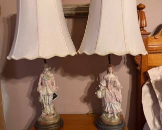 Pair of vintage MCM Italian porcelain figural table lamps features hand-painted 18th-century style aristocratic court or colonial couple figurines set on dark circular bases with scalloped fabric shades. Approx. 35h x 7" w base. $