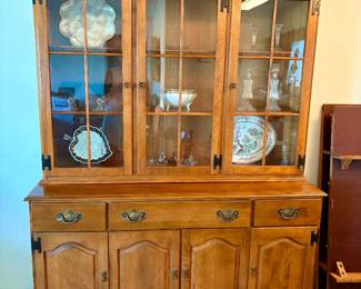 This beautiful vintage two-piece solid maple china cabinet and buffet hutch by Ethan Allen features a warm nutmeg finish, glass-paneled upper display doors, and classic brass-tone hardware.