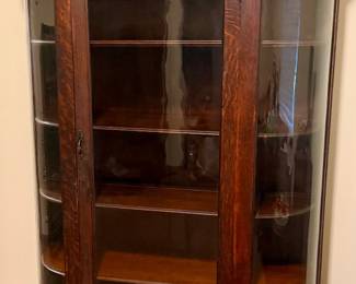 Antique bow-front curio / china cabinet with curved glass side panels, glass front door, interior display shelves, and decorative carved crest. Beautiful wood grain and elegant period styling. Researched prior to sale.