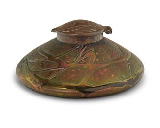 1027 An Art Nouveau bronze and art glass inkwell Late 19th/early 20th century Appears unmarked Possibly Loetz, with iridescent glass and hinged foliate-form bronze hinged lid opening to reveal a glass inkwell liner Height 2 3/8 in., diameter 5 3/8 in. Estimate: $300 - $500