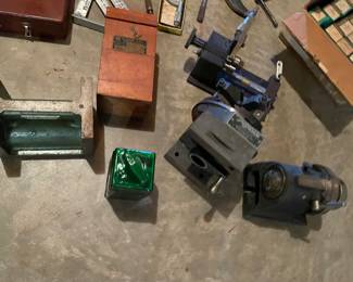 collection of vintage machine shop and machinist tooling, including a heavy cast-iron angle plate or V-block, a wooden gauge/tool box labeled "Gage Co.", a small green oil or fluid can, and precision workholding fixtures such as a 5C collet spin indexer/grinding fixture and a small milling/lathe attachment.