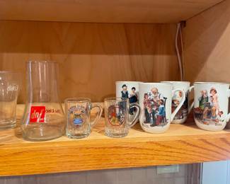 vintage glassware and porcelain mugs, including a Coca-Pele glass, a 7-Up glass, small commemorative glass mugs, and several Norman Rockwell Museum Collection porcelain coffee mugs featuring classic Americana artwork