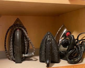 vintage electric clothes irons spanning different eras: a mid-century black-bodied iron with a patterned cord on the left, a heavy cast-iron sad iron in the middle, and a chrome-and-black Westinghouse steam/dry iron with a red temperature dial on the right.