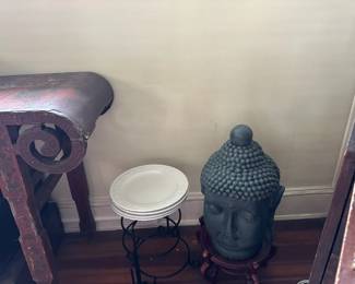 Decorative Buddha head statue, stacked white china plates, and carved console table