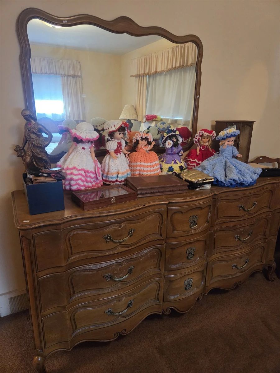 French Provincial nine-drawer triple dresser in a fruitwood finish with serpentine drawer fronts, brass pulls, and matching scalloped-frame mirror. The top displays a collection of handmade crochet-dressed bed dolls in Southern belle style, a bronze-finish figural statue, wooden display cases, and a small jewelry armoire.