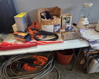 Garage table of household goods including an AT&T Trimline telephone in box, Sunbeam Mixmaster hand mixer in original box, Black and Decker blender with glass jar, Amerimax Flex-Elbow downspout elbow, orange extension cord, vacuum hose, golf balls and cap, and paper doilies; Black and Decker leaf blower and hedge trimmer with heavy garden hose below.