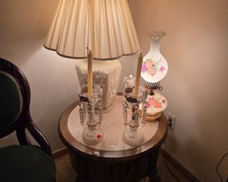 Round wood side table topped with a brass table lamp and a hand-painted floral milk glass lamp.