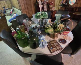 Mid-century modern dinette set with a white laminate oval table and molded scoop chairs upholstered in dark tortoise vinyl, shown loaded with smalls including two green glass demijohn jugs, pressed and cut glass vases, a jadeite-green vase, art glass, faux florals, baskets, and decorative trays. The set has clean atomic-era lines and sits ready for a retro kitchen.