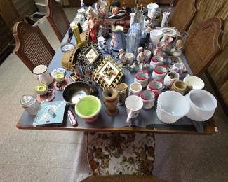 Dining table covered with collectible smalls including milk glass compotes and bowls, a set of Santa Claus mugs with red trim, figural salt and pepper sets, ornate gold-tone framed pictures, wicker wall pockets, ceramic candleholders, souvenir plates, and a brass bowl. Cane-back dining chairs with arched frames surround the table.