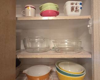 Cabinet of vintage Pyrex and ovenware including a Butterfly Gold Cinderella casserole, primary yellow and turquoise nesting mixing bowls, two white refrigerator dishes with Early American brown motif, clear glass casseroles with lids, and melamine bowls in lime and pink.