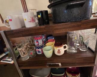 Three-tier woodgrain rolling serving cart loaded with collectible glassware and mugs, including Apollo moon landing commemorative glasses, character and advertising mugs, and beer steins. Two black speckled enamelware roasting pans sit on top, with stacks of colorful melamine square plates on the lower shelf.