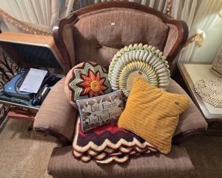 Wood-trimmed wingback armchair in taupe upholstery displaying handmade crochet pieces: a raised orange flower round pillow, a ruffled pastel round pillow, a gold corduroy pillow, an Old Friends are The Best Friends needlepoint pillow, and a scalloped chevron afghan.