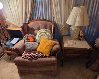 Brown wingback armchair with decorative throw pillows, paired with a wood side table and ceramic table lamp.