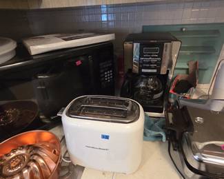 Small appliance grouping with a white Hamilton Beach two-slice toaster, a Magic Chef black microwave, and a programmable 12-cup drip coffee maker with glass carafe. A boxed hand mixer and glass storage pieces sit on the shelf above.
