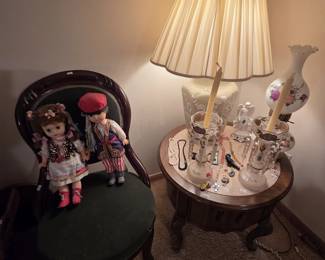 Victorian carved mahogany parlor chair with green velvet upholstery seating a pair of dolls in traditional Polish folk costume. The round French Provincial lamp table beside it holds a pair of white cased glass mantle lusters with hand-painted decoration and crystal prisms, a hand-painted hurricane lamp, an angel figurine, and assorted costume jewelry.