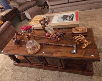 Wood coffee table with a lower storage cabinet and carved feet, topped with an assortment of decorative art objects including a gilded hand sculpture.