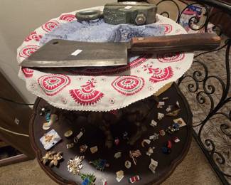 Vintage butcher cleaver with carbon steel blade and wood handle, a cast iron Mo-Clamp auto body pulling clamp, an embroidered folk art doily, and a tiered wood table spread with costume jewelry brooches and lapel pins including Christmas tree pins, American flag pins, and military ribbon bars.