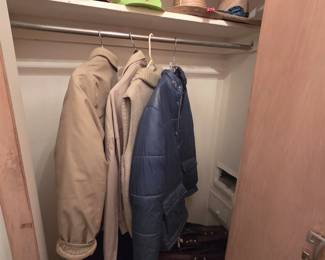 Closet of vintage mens outerwear including a khaki knit-collar jacket, a tan zip jacket, and a blue quilted parka, with a shelf of snapback and trucker caps, straw hats, plus boots and luggage below.