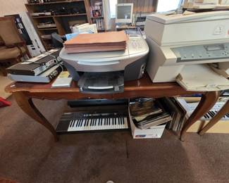 French Provincial console table with cabriole legs holding an HP Photosmart 2610 all-in-one printer and a Xerox WorkCentre copier, with a Casio Casiotone CT-360 keyboard, a second full-size keyboard, DVD players, and a box of vinyl records and sheet music below.