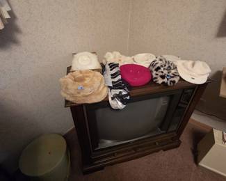 RCA wood console television displaying a collection of vintage ladies hats including faux fur styles, a fuchsia wool beret, a leopard print hat, and a boxed white fur hat, along with a piano keyboard print chiffon scarf and an oval hat box on the floor.