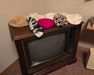 Vintage wood console television with a CRT screen, topped with several decorative hats.