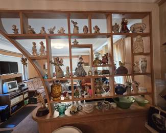Full view of a built-in mid-century blond wood room divider filled with porcelain angels, boy and girl figurines, bud vases, an amber glass fairy lamp, Polish costumed dolls, a snow globe music box, teapots, vases, and green depression-style glass bowls along the base.