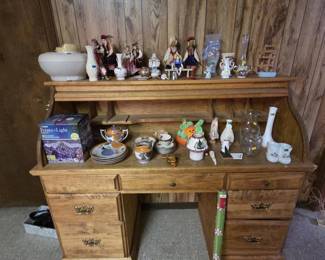 Maple roll-top style kneehole desk with seven drawers and brass bail pulls, topped with a collection of Polish folk art dolls in traditional Krakow costume, hand-painted ornaments, porcelain vases, a Lenox-style ewer, hand-painted Japanese lusterware plates and tea pieces, cut glass, milk glass, and a boxed Points of Light projection lamp.
