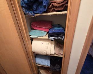 Linen closet stocked with handmade crocheted afghans in blue and brown tones, bath rugs, towels, a cream quilted coverlet, embroidered table linens, bed sheets, a round braided rug, and a plastic step stool.