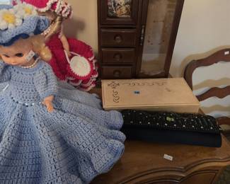 Vintage wooden jewelry armoire with drawers, a glass door, and a Victorian courting-scene print panel, shown with a handmade crochet-dressed bed doll in powder blue with daisy bonnet, a cream hinged dresser box with cutwork detail, and a black beaded evening clutch.