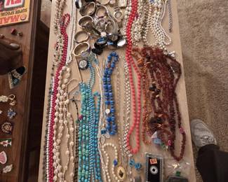 Large costume jewelry lot laid out on a table: Baltic amber chip necklaces, turquoise-colored and blue art glass bead necklaces, coral-red and milk glass beads, cowrie shell leis, faux pearls, rosaries, a rhinestone American flag brooch, western belt buckles, and a collection of vintage ladies and mens wristwatches with stretch bands.