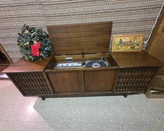Mid-century walnut stereo console with lift top revealing an AM-FM tuner and record changer turntable, flanked by lattice-front speaker grilles on a raised leg base. Shown with a decorated holiday wreath and a carved and painted wooden folk art wall plaque of an alpine chalet.