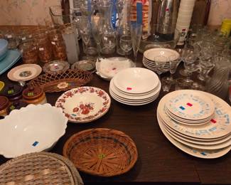 Table with a boxed Farberware set, stacked dinner plates, wicker placemats, and assorted glassware.