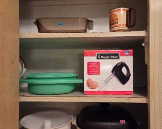 Cabinet shelf with a boxed Magic Chef hand mixer, a green baking dish, and Corningware-style covered pots.