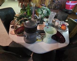 Mid-century laminate dinette table with six black vinyl swivel chairs, topped with a lot of glassware including a crystal vase, a green two-handled art glass vase, a ruby glass shell dish, pressed glass compote, rose-pattern china cups and saucers, green glass demijohn bottles, a ceramic planter bassinet, and a portable electric heater.