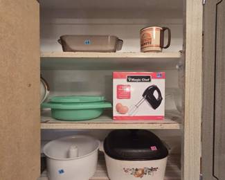 Cabinet shelf lot including a Magic Chef 5-speed hand mixer new in box, a smoke glass loaf pan, a Dawn Donuts advertising mug, a jadeite green Tupperware steamer bowl, a white ceramic angel food cake pan, and a Corning Ware style covered casserole with fruit motif and black lid.