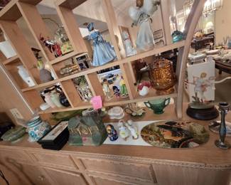 Mid-century blond wood room divider shelf displaying collectible figurines including a porcelain lady in blue, an angel figurine, an amber glass fairy lamp, a Russian matryoshka nesting doll, a Polish costumed doll, a cast cottage clock, a hand-painted portrait plaque, and a porcelain ballerina on a wood base.