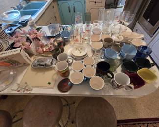 Kitchen table spread of glassware and china including stemware, wine glasses, tumblers, teacups, saucers, melamine cups in blue and beige, a serving caddy in box, a chrome covered butter dish, smoke glass tumblers, and a floral luncheon plate set.