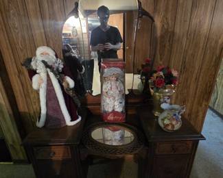 Antique early 20th century vanity in walnut finish with dramatic carved-crest tri-fold style mirror, lower center well, flanking drawer pedestals on casters, displayed with a plush Father Christmas figure, a boxed Century Collection porcelain doll, an oval woven-framed mirror, and a hand-painted pitcher.