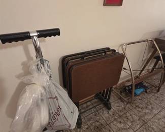 Set of vintage vinyl-top folding TV tray tables on their original rolling storage rack, alongside a chrome utility cart and a brass finish rolling luggage rack. A framed mid-century chalkware relief of a fisherman hangs above.
