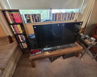 Vizio flat-screen smart TV with remote displayed on an oak coffee table, alongside an LED digital clock, vintage speakers, and shelves of VHS tapes including a Polish Army collection, Cleopatra, and other classic films.