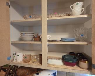 Upper cabinet of vintage glass including pink depression glass bowls and creamer, white milk glass hobnail sugar and creamer set, clear swirl glass dishes, ivory stoneware bowls, powder blue saucers, a covered butter dish, stoneware crock mugs, and small casserole dishes in green and burgundy.