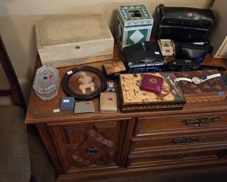 Dresser-top collection of smalls including a Bell and Howell leather camera bag, a Chinon 35FA Super 35mm camera in box, a Canon point-and-shoot camera, a vintage travel alarm clock, a cream jewelry chest, a crystal covered dish, a round framed antique baby photograph, ladies cigarette cases and compacts, a wristwatch, and two carved wood-burned folk art boxes.