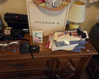 Woven straw ladies hat decorated with deep pink chrysanthemum petals and a large sheer butterfly, displayed on a Hudsons department store box. Also shown are an Optimus pocket radio, a shell-form travel case, a floral anniversary keepsake book, a ladies wristwatch, and a stack of new-in-package hosiery and knee highs.