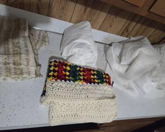 Handmade textiles including a cream crocheted afghan with a bold multicolor granny-stitch band, a woven beige-and-cream knit top, and folded white sheers and linens.
